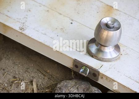 Nahansicht: Rustikale weiße Holztürverkleidung mit silbernem Metallknopf und Verriegelungsmechaniker über schmutzigem Boden, die verlassene Elemente darstellen Stockfoto