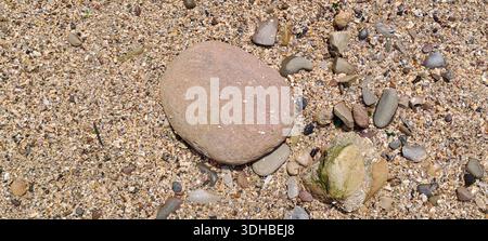 Glatter rötlicher Stein hebt sich zwischen kleineren Felsen und Kieselsteinen an einem Sandstrand hervor und schafft eine ruhige Küstenlandschaft Stockfoto