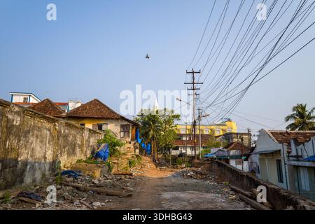Fort Kochi, Indien - 16. März 2023: In Fort Kochi wird eine Wohnstraße mit verstreuten Trümmern und Schutt gezeigt, mit alten Häusern mit Ziegeldächern, Freileitungen, Versorgungsmasten und einem Vogel, der am Himmel fliegt. Stockfoto