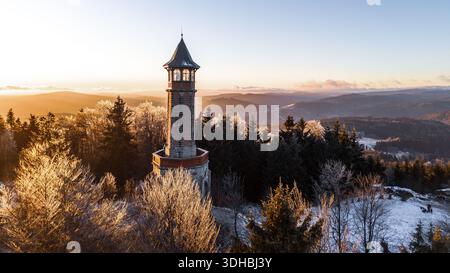Der Stepanka Aussichtsturm steht hoch im Isergebirge, wenn die Sonne untergeht. Die Landschaft mit Bäumen und leichtem Schnee bietet einen klaren Blick auf die umliegenden Hügel und Täler. Stockfoto