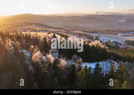 Der Aussichtsturm im Isergebirge steht bei Sonnenuntergang hoch. Frost bedeckt die Bäume und schafft eine Winterszene. Die umliegenden Hügel und Felder sind unter dem schwindenden Licht sichtbar. Stockfoto
