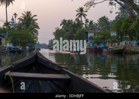 Alappuzha, Indien - 7. März 2023: Mehrere Menschen werden auf Holzbooten entlang eines Kanals gesehen, der von Palmen, Häusern und einer Moschee gesäumt ist. Stockfoto
