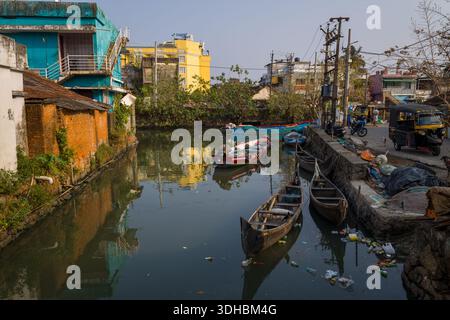 Fort Kochi, Indien - 16. März 2023: Ein Kanal im Zentrum von Fort Kochi ist gesäumt von Holzbooten, vertäuten Fischereifahrzeugen und Trümmern, die im Wasser schwimmen. Farbenfrohe Gebäude, Elektromasten, Auto-Rikschas und Motorräder sind entlang der Straße sichtbar. Stockfoto