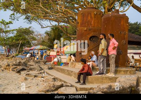 Fort Kochi, Indien - 16. März 2023: Mehrere Personen sitzen und stehen neben zwei großen, rostfreien Dampfschiffkesseln auf einer Betonplattform in der Nähe des felsigen Strandes, mit Marktständen und Bäumen im Hintergrund. Stockfoto