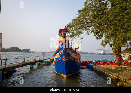 Fort Kochi, Indien - 16. März 2023: Ein bunt bemaltes Fischerboot liegt am Kai an, mit mehreren Leuten, die neben roten Fischernetzen unter einem großen Baum am Ufer sitzen, und Hafenkranichen, die im Hintergrund über dem Wasser sichtbar sind. Stockfoto