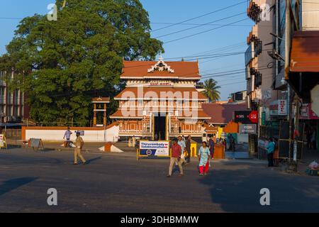 Thrissur, Indien - 12. März 2023: Menschen gehen zu Fuß und Polizeibeamte stehen vor dem Eingang des Paramekkavu Bhagavathi Tempels, mit Schildern, Geschäftsgebäuden und einem großen Baum, der im Hintergrund zu sehen ist. Stockfoto