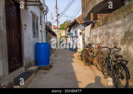 Fort Kochi, Indien - 16. März 2023: Eine enge Gasse, gesäumt von verwitterten Wohngebäuden, mehreren geparkten Fahrrädern, einem großen blauen Wasserfass, einer Wäscherei, die an einer Wand hängt, und bei der Nachmittagssonne sichtbare Stromleitungen. Stockfoto