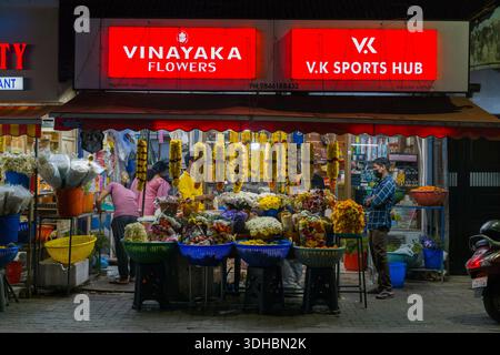 Alappuzha, Indien - 8. März 2023: Mehrere Menschen werden nachts vor dem Vinayaka Flowers Shop gesehen, in dem Blumengirlanden, Blumensträuße und Blumenkörbe unter beleuchteten Schildern ausgestellt werden, wobei ein Mann steht und andere Blumen arrangieren. Stockfoto