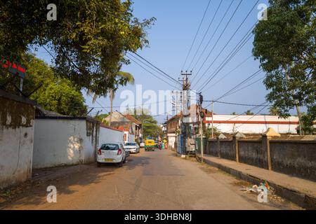 Fort Kochi, Indien - 16. März 2023: Eine Straßenszene in Fort Kochi zeigt geparkte Autos, eine Auto-Rikscha, Freileitungen, Grenzmauern und Bäume, die beide Seiten der Straße bei klarem Tageslicht säumen. Stockfoto