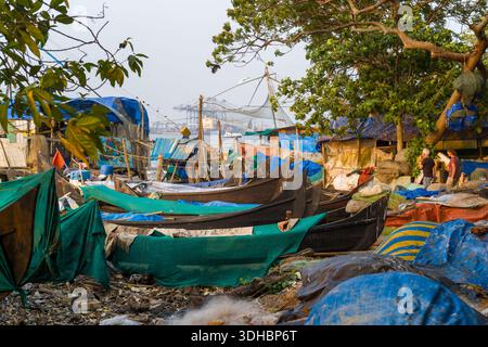 Fort Kochi, Indien - 16. März 2023: Mehrere Holzfischboote liegen entlang einer überdachten Uferpromenade mit provisorischen Unterkünften, die mit Planen, Fischernetzen, Trümmern und ein paar Menschen in der Nähe der Boote bedeckt sind, während Hafenkrane im Hintergrund über dem Wasser sichtbar sind. Stockfoto