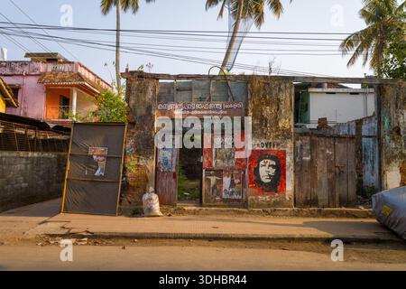 Fort Kochi, Indien - 16. März 2023: Die Fassade eines verlassenen Gebäudes zeigt verwitterte Wände, politische Plakate, ein Wandbild von Che Guevara und Schilder in Malayalam, mit Palmen und Wohngebäuden im Hintergrund. Stockfoto