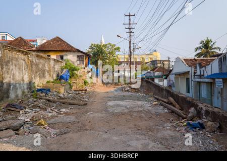 Fort Kochi, Indien - 16. März 2023: Eine unbefestigte Straße, die mit verstreuten Trümmern, Schutt und Bauabfällen gesäumt ist, wird von alten Häusern mit Ziegeldächern, verwitterten Wänden und Überkopfkabeln begrenzt, mit Versorgungsmasten und einer Palme, die im Hintergrund sichtbar sind. Stockfoto