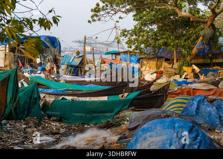 Fort Kochi, Indien - 16. März 2023: Mehrere Holzfischboote liegen entlang einer überdachten Uferpromenade mit provisorischen Unterkünften, die mit Planen, Fischernetzen und Trümmern bedeckt sind, während einige Menschen in der Nähe der Boote stehen und Hafenkrane im Hintergrund über dem Wasser sichtbar sind. Stockfoto