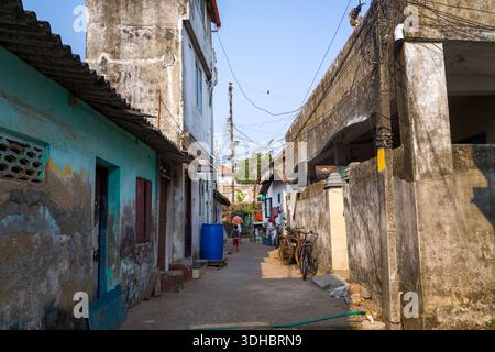 Fort Kochi, Indien - 16. März 2023: Eine enge Gasse, gesäumt von verwitterten Wohngebäuden, Fahrrädern, einem blauen Wasserfass und sichtbaren elektrischen Leitungen über dem Kopf, wobei eine Person in der Ferne läuft. Stockfoto