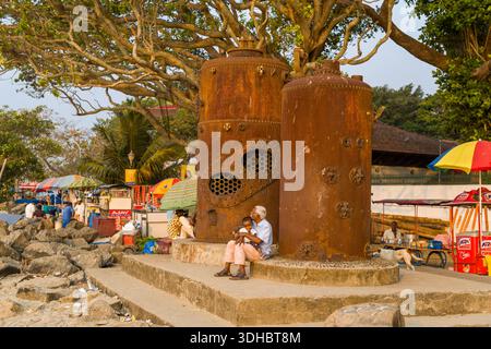 Fort Kochi, Indien - 16. März 2023: Mehrere Personen sitzen und stehen neben zwei großen, rostfreien Dampfschiffkesseln auf einer Betonplattform in der Nähe des felsigen Strandes, mit Marktständen und Bäumen im Hintergrund. Stockfoto