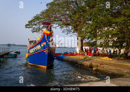 Fort Kochi, Indien - 16. März 2023: Das Fischerboot Badriyyah liegt am Kai an, mit einer Gruppe von Menschen, die sich in der Nähe von roten und gelben Fischernetzen unter großen Bäumen am Ufer versammelt haben, und Hafenkranichen, die im Hintergrund über dem Wasser sichtbar sind. Stockfoto