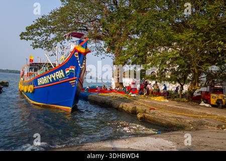 Fort Kochi, Indien - 16. März 2023: Das Fischerboot Badriyyah liegt am Kai an, mit einer Gruppe von Menschen, die sich in der Nähe roter Fischernetze unter großen Bäumen am Ufer versammelt haben, und Hafenkranichen, die im Hintergrund über dem Wasser sichtbar sind. Stockfoto