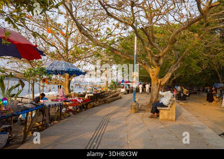Fort Kochi, Indien - 16. März 2023: Mehrere Straßenverkäufer stellen Waren auf Tischen unter großen Sonnenschirmen neben einem gepflasterten Gehweg, der von blattlosen und belaubten Bäumen gesäumt ist, aus. Die Leute sitzen auf Bänken und laufen in der Nähe des Ufers. Stockfoto