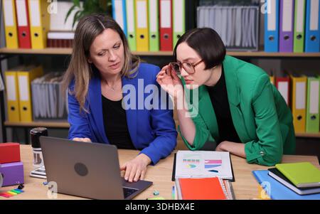 Zwei Profis arbeiten an einem Laptop in einem lebhaften Büro mit farbenfrohen Ordnern und Dokumenten an einem geschäftigen Arbeitstag zusammen Stockfoto