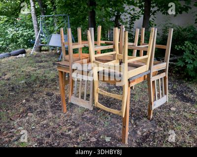 Mehrere Holzstühle stapelten sich kopfüber auf einem hölzernen Esstisch in einem Gartenbereich mit Bäumen und einer Gartenschaukel. Stockfoto