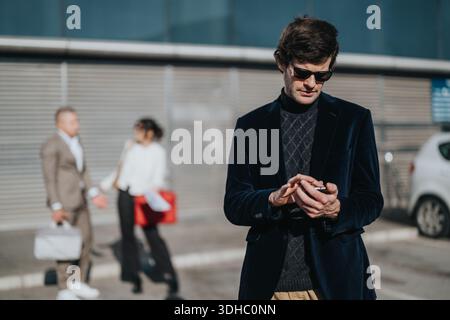 Ein stilvoller Mann überprüft sein Handy, während Kollegen im Hintergrund in der städtischen Geschäftsszene chatten Stockfoto