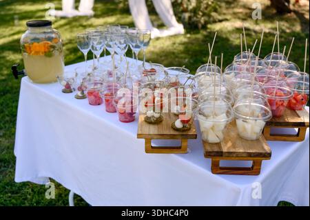 Ein Buffet mit Snacks in Plastikbechern und Limonade. Portioniertes Obst und Mozzarella mit Tomaten auf einer weißen Tischdecke für Catering im Freien Stockfoto