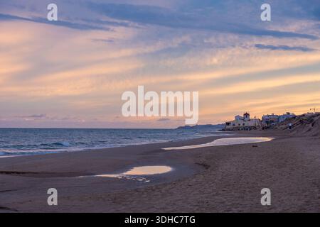 Ein ruhiger Strand bei Sonnenuntergang mit weichen pastellfarbenen Wolken über dem Meer. Eine ferne Küstenstadt liegt neben Dünen. Nasser Sand reflektiert das Licht und erzeugt eine Erbse Stockfoto