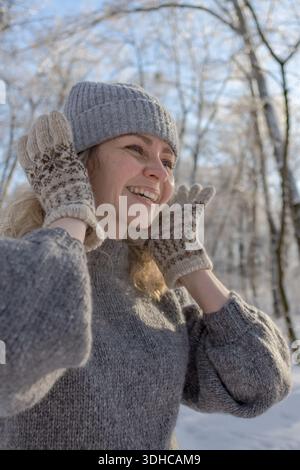 Ziemlich glückliches Mädchen in einem warmen grauen Hut und Pullover. Strickpullover mit Elchmuster. Hände in warmen Fäustlingen. Authentische ukrainische handgefertigte Kleidung. Stockfoto