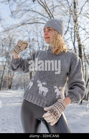 Ziemlich glückliches Mädchen in einem warmen grauen Hut und Pullover. Strickpullover mit Elchmuster. Hände in warmen Fäustlingen. Authentische ukrainische handgefertigte Kleidung. Stockfoto