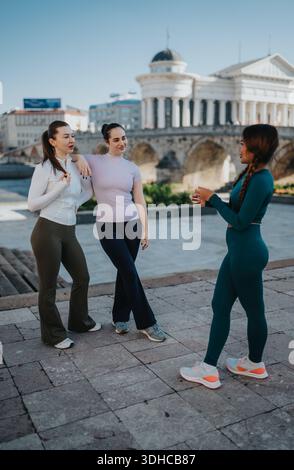 Drei Freunde unterhalten sich und posieren im Freien in sportlichen Outfits in der Nähe eines historischen Flussufers und eines großen Gebäudes Stockfoto