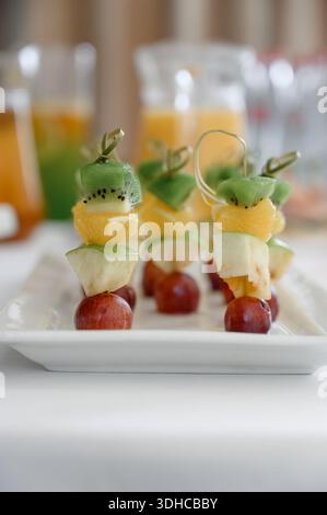 Dessertspieße mit geschnittenen tropischen Früchten auf einem festlichen Tisch. Frische Obstkanapees aus Kiwi, Orange, Apfel und Trauben auf einem weißen Buffet-Tablett Stockfoto