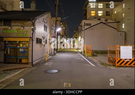 Kyoto, Japan - 15. Oktober 2023: Eine ruhige Straße bei Nacht zeigt eine Apotheke mit beleuchteten Schildern auf der linken Seite, Baubarrieren und Schildern auf der rechten Seite, Freileitungen und Wohngebäude im Hintergrund. Stockfoto