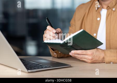 Ein Mann, der Notizen in einem grünen Tagebuch macht, Ideen festnimmt und Aufgaben mit einem Stift plant, mit einem Laptop an einem Holzschreibtisch sitzt und sich auf Organisation und Produktivität konzentriert Stockfoto