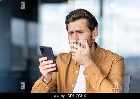 Junger bärtiger Mann in einem modernen Büro, der verstört aussah, während er eine beunruhigende Benachrichtigung auf seinem Smartphone las, während er sich während der Fernarbeit Sorgen über sein Gesicht machte Stockfoto