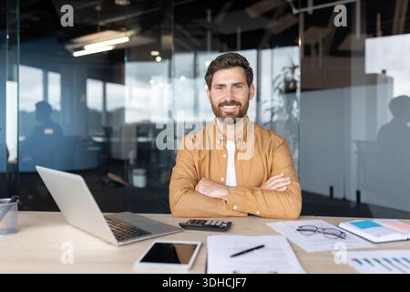 Junger männlicher Unternehmer, der mit Laptop und Dokumenten am Schreibtisch sitzt, in die Kamera lächelt und Erfolge, Startup und berufliche Leistungen schildert Stockfoto