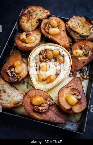 Im Ofen gebackener Camembert-Käse mit karamelisierten glasierten Birnen, Walnüssen und Trauben sowie Baguette auf rustikalem Metallteller Stockfoto
