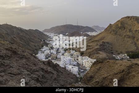 Blick aus der Vogelperspektive auf weiße Gebäude inmitten zerklüfteter, sonnenverwöhnter Berge, im Kontrast zum klaren Himmel, Muscat, Muscat Governorate, Oman. Stockfoto