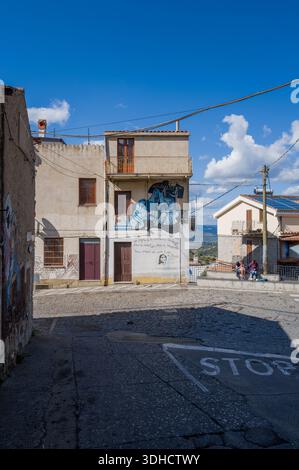 Orgosolo, Italien – 25. September 2023: An der Fassade eines Wohnhauses an einer Straßenkreuzung mit Kopfsteinpflaster ist ein Wandbild mit blauen abstrakten menschlichen Figuren und handgeschriebenem italienischem Text gemalt. Stockfoto