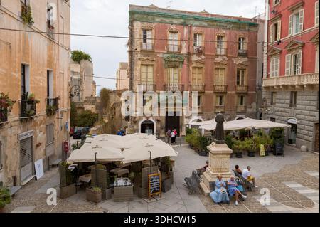 Cagliari, Italien - 20. September 2023: Auf einem kleinen Stadtplatz befindet sich eine Statue auf einem Sockel, auf Bänken sitzende Menschen, ein Café im Freien unter Sonnenschirmen und die umliegenden historischen mehrstöckigen Gebäude mit verwitterten Fassaden und Balkonen. Stockfoto
