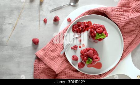 Zwei Gläser leuchtend roter Himbeerpudding werden auf einem weißen Teller mit frischem Obst und rosa Waffelwäsche serviert. Natürliches Licht hebt den Glanz hervor Stockfoto