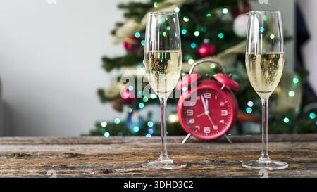 Zwei Sektgläser sitzen auf einem Holztisch. In der Nähe befindet sich eine rote Uhr, die eine festliche Kulisse mit einem mit Lichtern und Ornamenten verzierten Weihnachtsbaum zeigt Stockfoto