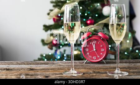 Zwei Sektgläser sitzen auf einem Holztisch neben einem roten Wecker vor einem Weihnachtsbaum mit Lichtern und Dekorationen. Die Szene zeigt einen holi Stockfoto