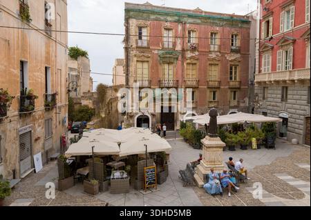 Cagliari, Italien - 20. September 2023: Auf einem kleinen Stadtplatz befindet sich eine Statue auf einem Sockel, auf Bänken sitzende Menschen, ein Café im Freien unter Sonnenschirmen und die umliegenden historischen mehrstöckigen Gebäude mit verwitterten Fassaden und Balkonen. Stockfoto