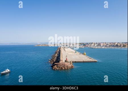 Porto Torres, Italien - 29. September 2023: Die lange Mole erstreckt sich bis ins Meer am Eingang zum Hafen, mit einem kleinen Boot am Ende und dem Stadtgebiet von Porto Torres, das im Hintergrund unter einem klaren blauen Himmel zu sehen ist. Stockfoto
