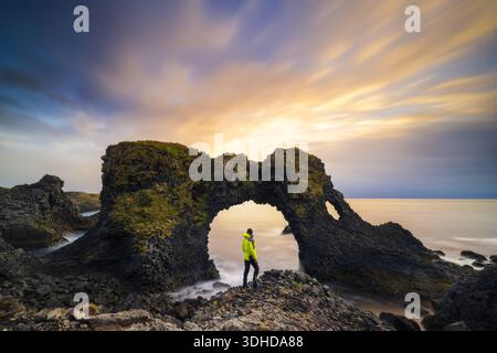 Der Blick auf eine Einzelfigur steht unter einem Natursteinbogen, während die Sonne den Himmel in leuchtenden Farben färbt und eine atemberaubende Szene schafft: Arnarstapi, S Stockfoto