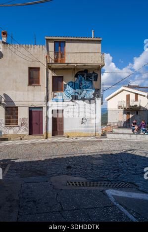 Orgosolo, Italien – 25. September 2023: An der Fassade eines Wohnhauses an einer Straßenkreuzung mit Kopfsteinpflaster ist ein Wandbild mit blauen abstrakten menschlichen Figuren und handgeschriebenem italienischem Text gemalt. Stockfoto