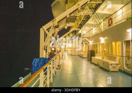 Unbekannter Ort - 13. September 2023: Das Bild zeigt das Außendeck einer Fähre bei Nacht mit Bänken, weißen Aufbewahrungsboxen mit der Aufschrift „DRILL“, einem mit blauer Plane bedeckten Objekt, einem Metallgeländer und einer sichtbaren Rettungsbootstruktur unter künstlicher Beleuchtung. Stockfoto