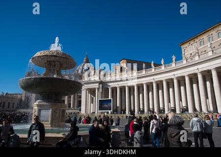 Touristen und Besucher stehen an einem sonnigen Tag auf dem Petersplatz im Vatikan in Rom mit klarem blauen Himmel und Brunnen im Vordergrund. Stockfoto