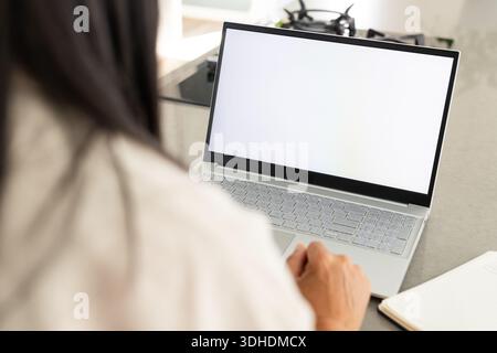 Der silberne Laptop sitzt offen auf einer grauen Arbeitsplatte neben dem Fenster, das Notebook und der Gasherd sind sichtbar Stockfoto
