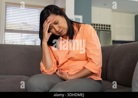 Asiatische Frau mittleren Alters, die zu Hause auf einer grauen Couch sitzt, in einem orangefarbenen Hemd, der Kopf und Bauch hält Stockfoto